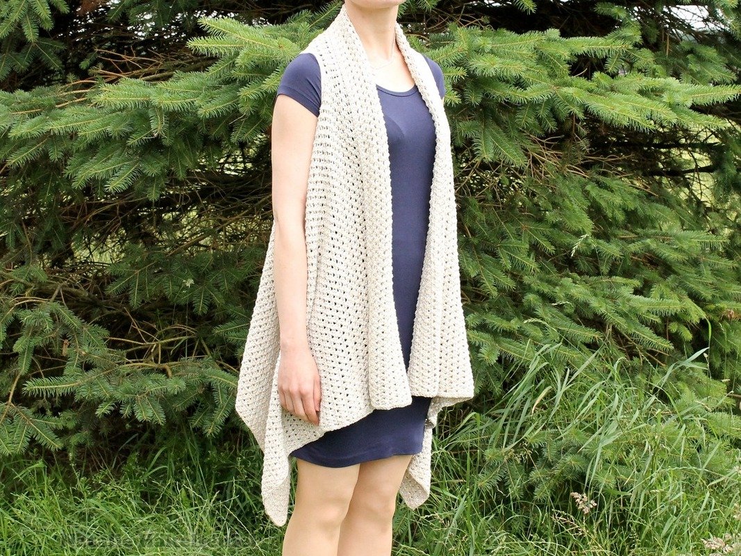 Person trägt helle, lange gehäkelte Weste über dunklem Kleid vor Tannenzweigen