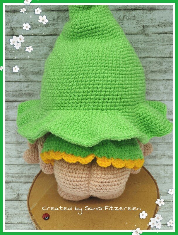 Rückansicht eines gehäkelten Amigurumi-Wichtels mit großem grünem Spitzhut, grüner Kleidung und gelbem Saum auf einer Holzscheibe.