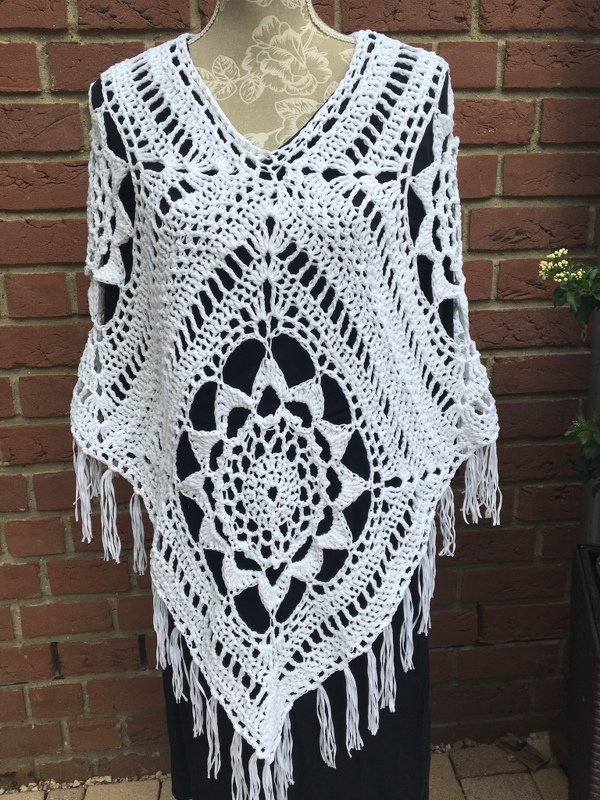 Weißer gehäkelter Damenponcho mit zentralem Mandala, V-Ausschnitt und Fransen auf Schneiderpuppe vor Backsteinwand
