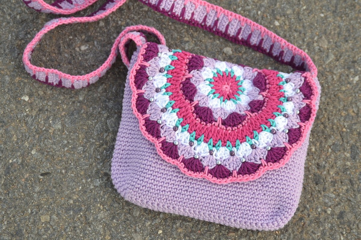 Eine gehäkelte Umhängetasche in Lila mit farbigem Mandala-Muster und Perlen, liegend auf einem grauen, steinigen Untergrund.