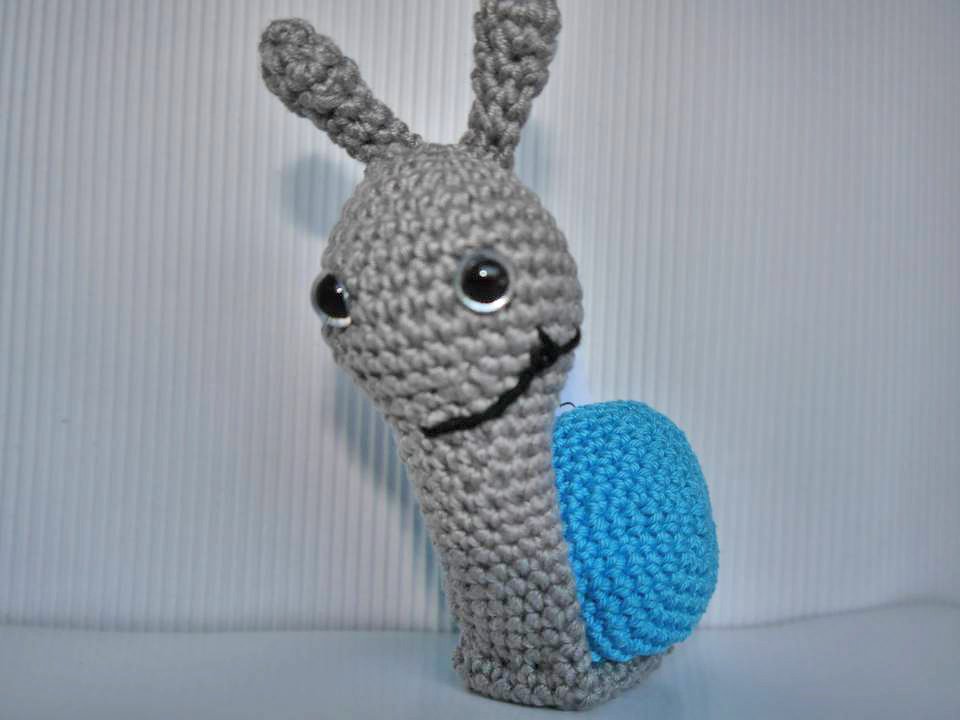 Gehäkelte graue Amigurumi‑Schnecke mit blauer Schale vor hellem Hintergrund