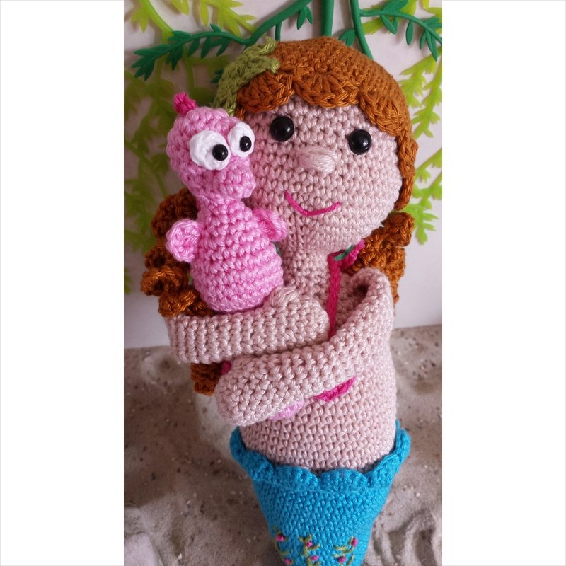 Gehäkelte Meerjungfrauenpuppe mit rötlich-braunen Haaren und blauem Schwanz, die ein rosa Seepferdchen hält.