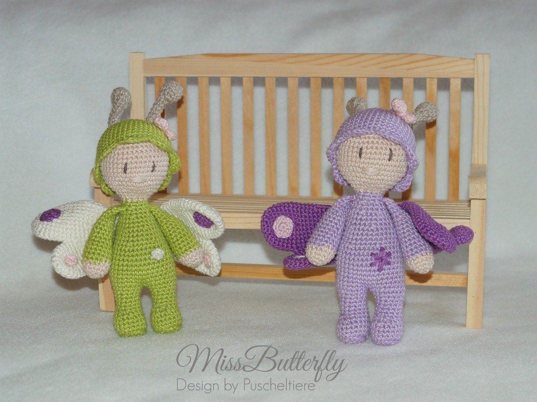Zwei gehäkelte Amigurumi-Schmetterlingspuppen, eine grüne und eine lila, vor einer Holzbank.