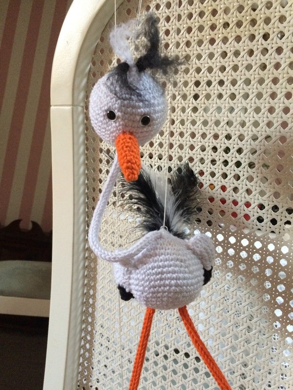 Gehäkeltes Amigurumi-Storch-Mobile in Weiß mit orangefarbenem Schnabel, langen Beinen und Feder-Akzenten, am Faden vor geflochtenem Hintergrund hängend.