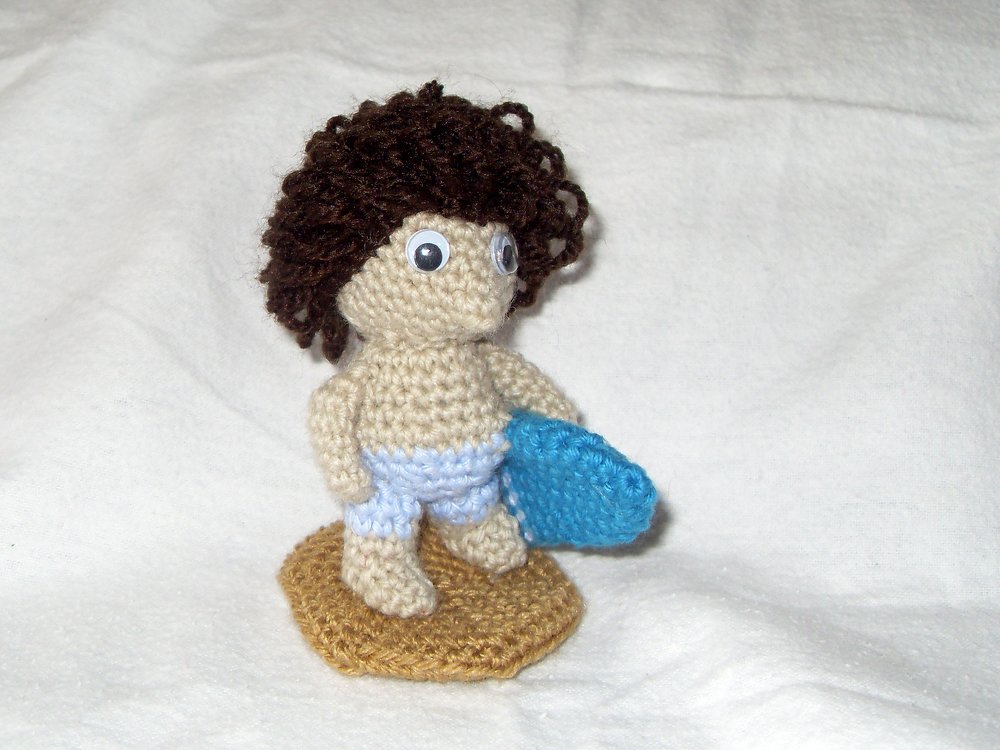 Gehäkelte Mini‑Puppe mit braunen Locken, blauer Badehose und blauem Surfbrett auf runder sandfarbener Basis