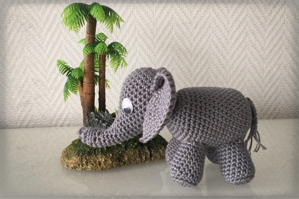 Grau gehäkelter Amigurumi-Elefant mit Wackelaugen neben Mini-Palmen