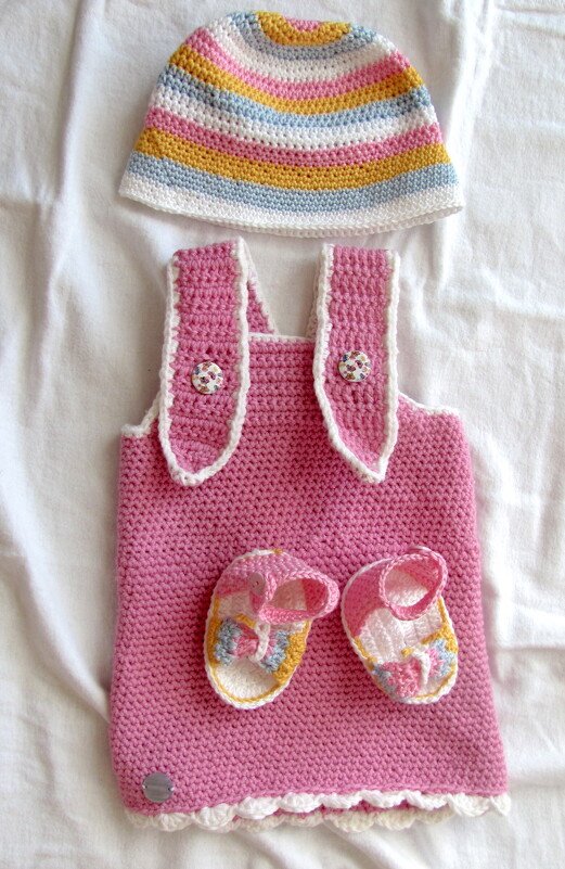Gehäkeltes rosa Baby-Trägerkleid mit gestreifter Mütze und Schühchen auf weißem Stoff