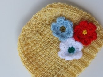 Gelbe Babymütze mit Rippenmuster und drei gehäkelten Blumen (blau, rot, weiß)