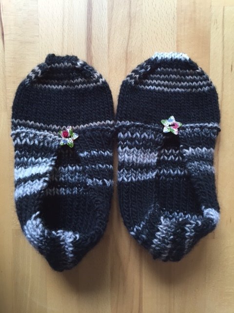 Paar schwarz-grauer gestrickter Hausschuhe mit Blumenknöpfen auf hellem Holz
