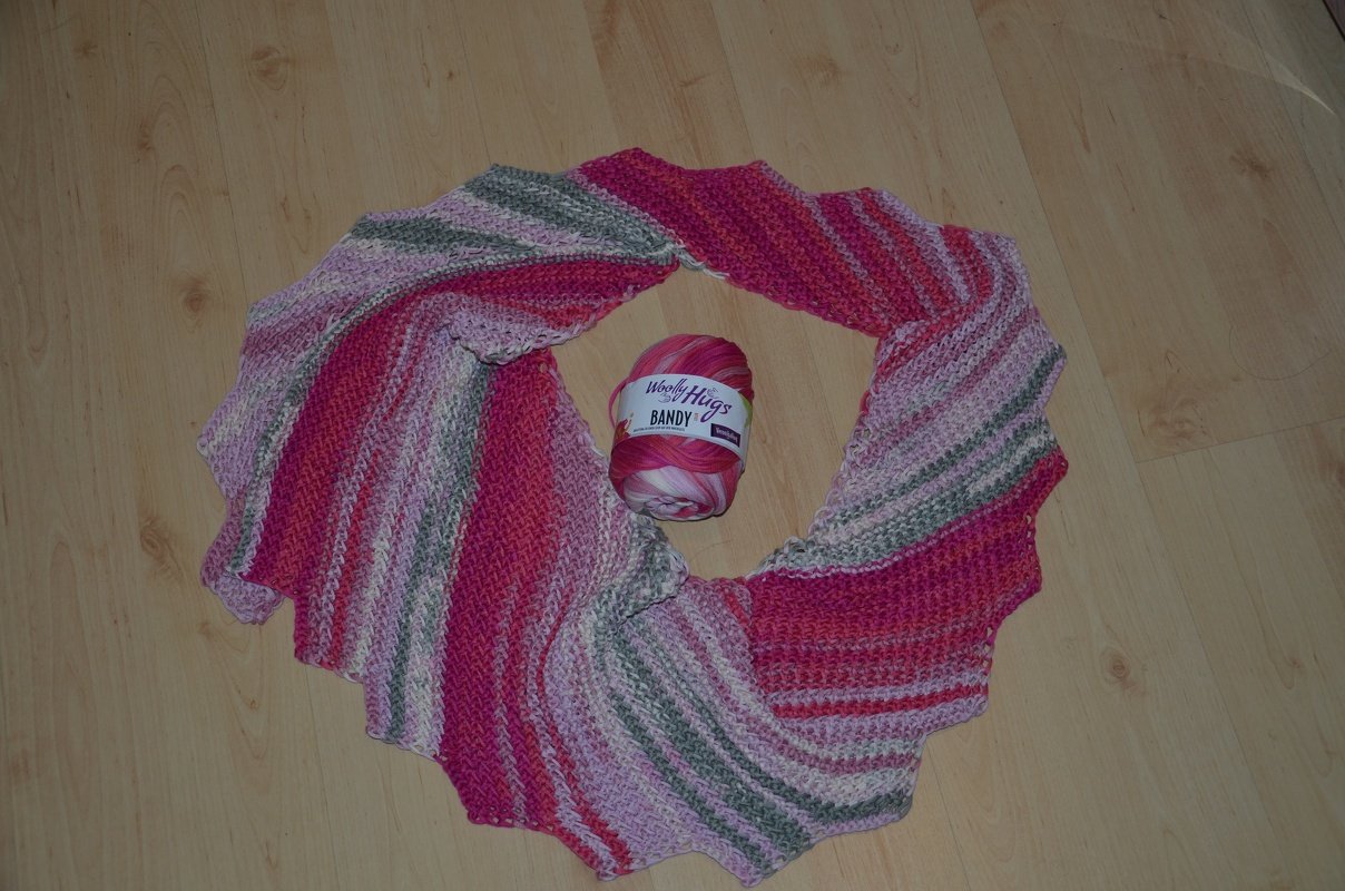 Gestrickter Drachenschwanz-Schal in Rosa, Lila, Grau und Weiß mit farblich passendem Wollknäuel auf hellem Holzboden.