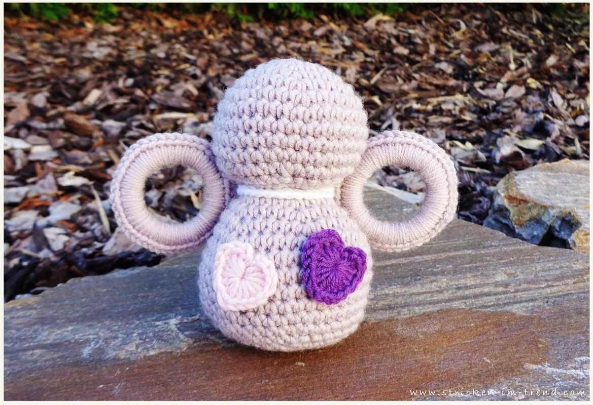 Gehäkelte lila Babyrassel mit zwei runden Greifringen und zwei Herzapplikationen auf einer Steinoberfläche