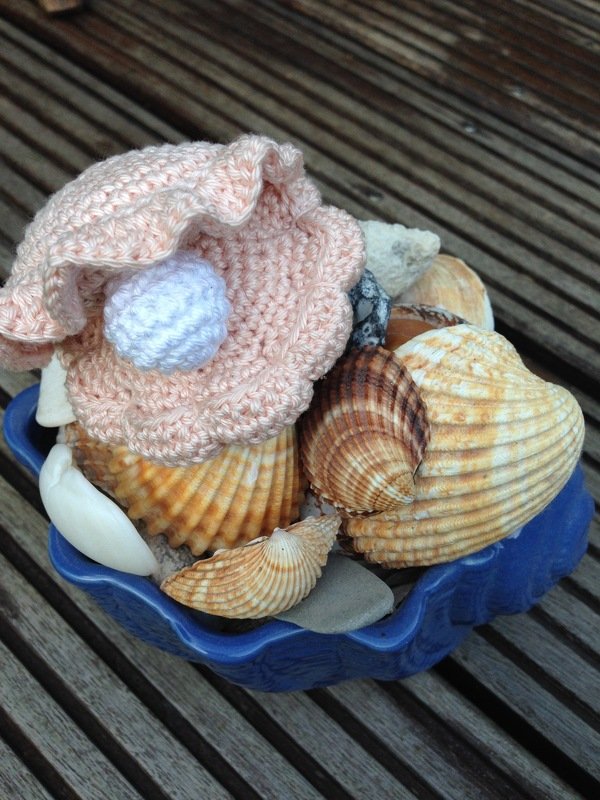 Gehäkelte rosa Muschel mit weißer Perle in einer blauen Schale voller Muscheln auf einem Holztisch.