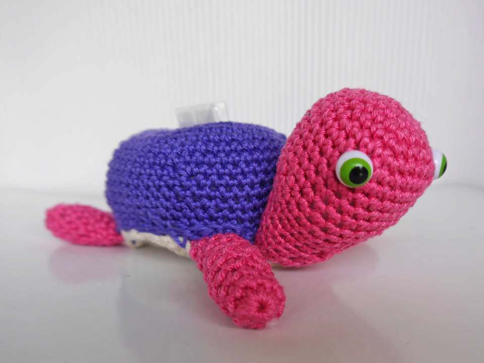 Gehäkelte Amigurumi‑Schildkröte mit rosa Kopf und Beinen, lila Panzer und großen grünen Augen auf hellem Untergrund.