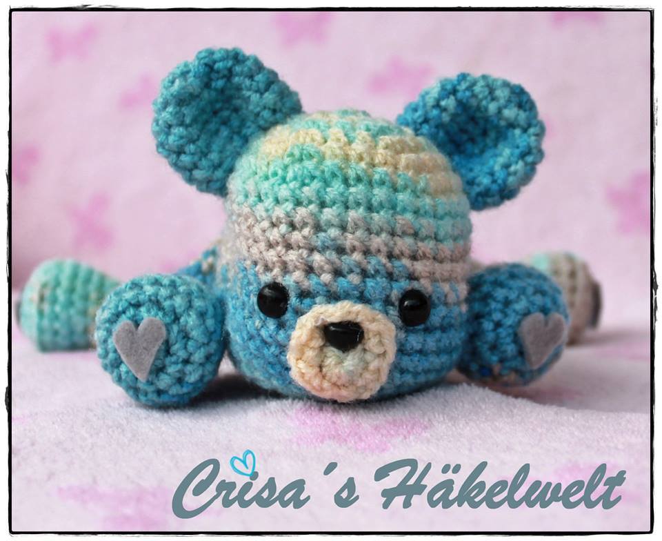Gehäkelter Amigurumi-Teddybär in Blau- und Türkistönen auf rosafarbenem Untergrund, mit grauen Herz‑Applikationen an den Pfoten.