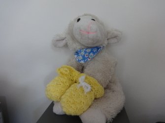 Weißes Stofflamm mit blauem Halstuch hält gelbe, gehäkelte Babyfäustlinge.