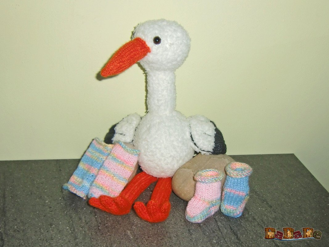 Gestrickter weißer Amigurumi-Storch mit orangefarbenem Schnabel und roten Beinen zwischen pastellgestreiften Babysocken