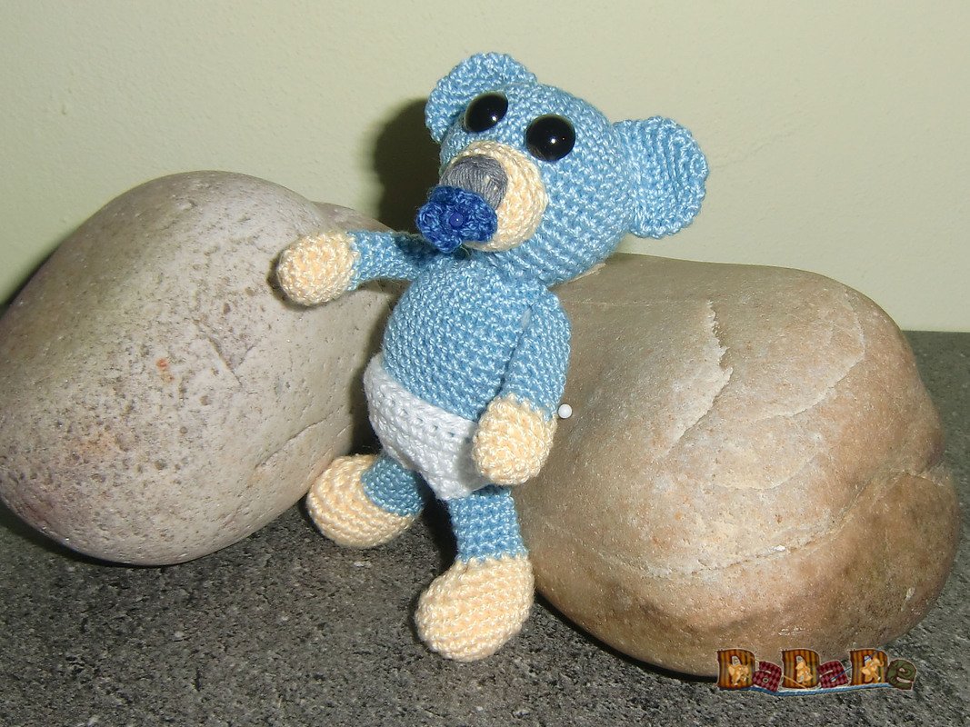 Hellblauer gehäkelter Amigurumi-Teddy mit weißer Windel und blauem Schnuller vor zwei Steinen