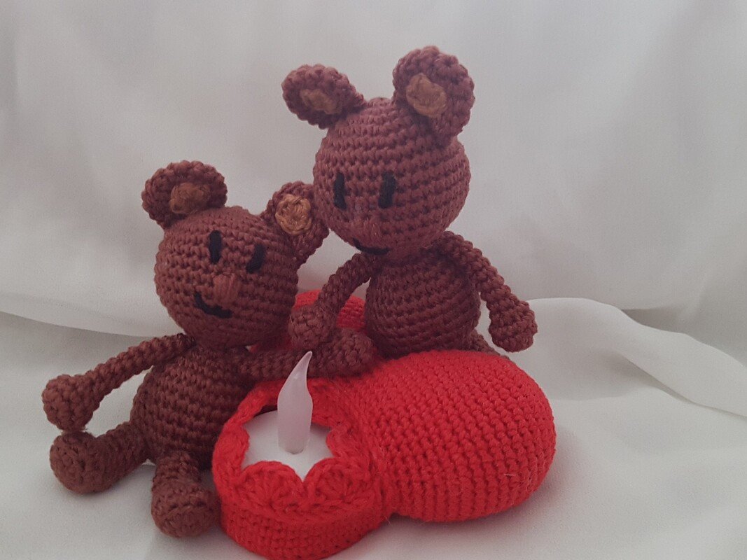 Zwei braune gehäkelte Amigurumi‑Bären neben rotem gehäkeltem Herz mit dekorativer Kerzenflamme