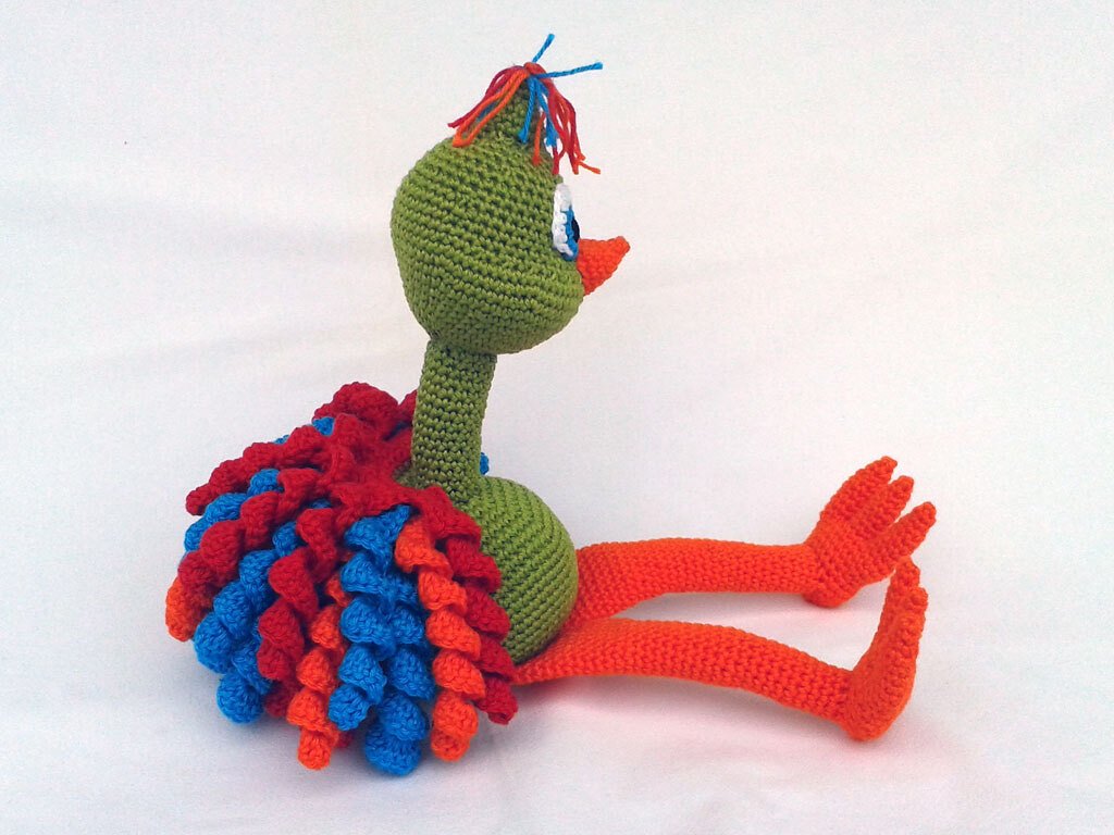 Gehäkelter Amigurumi-Strauß mit grünem Körper, bunten Rüschenfedern und orangefarbenen Beinen vor weißem Hintergrund.
