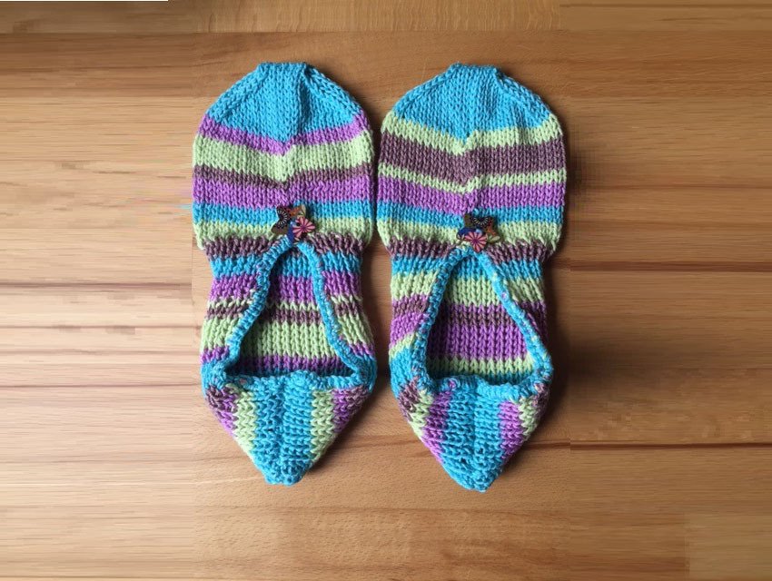 Gestrickte Damenpantoffeln mit bunten Streifen und kleinem blumenförmigem Knopf auf Holzboden.