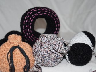 Mehrere gehäkelte Hundespielzeuge auf hellem Stoff: schwarzer Ring mit pinken Stichen, mehrfarbige Kugel, orangefarbener Knoten und schwarz-weißes Zylinderstück.