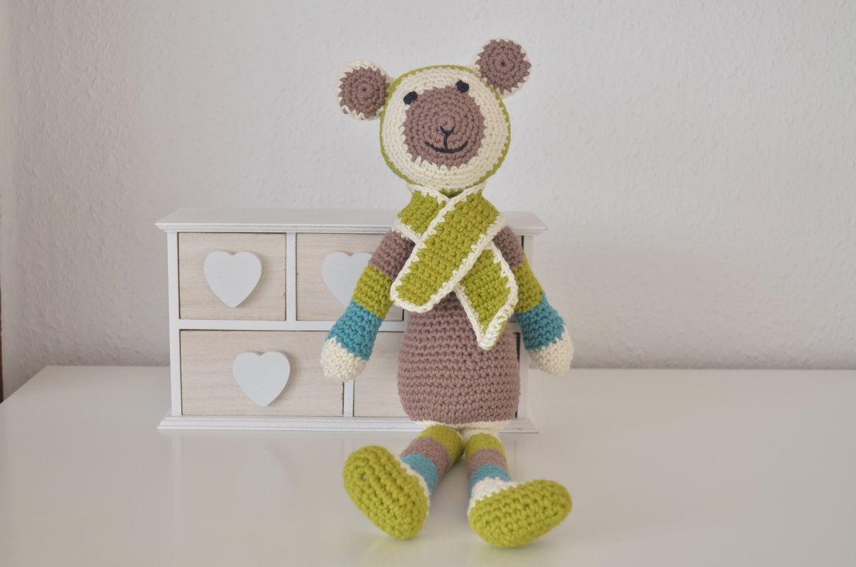 Gehäkelter Amigurumi-Teddybär mit braun-beigem Körper, gestreiften Gliedmaßen und hellgrünem Schal, sitzend vor einer weißen Kommode.