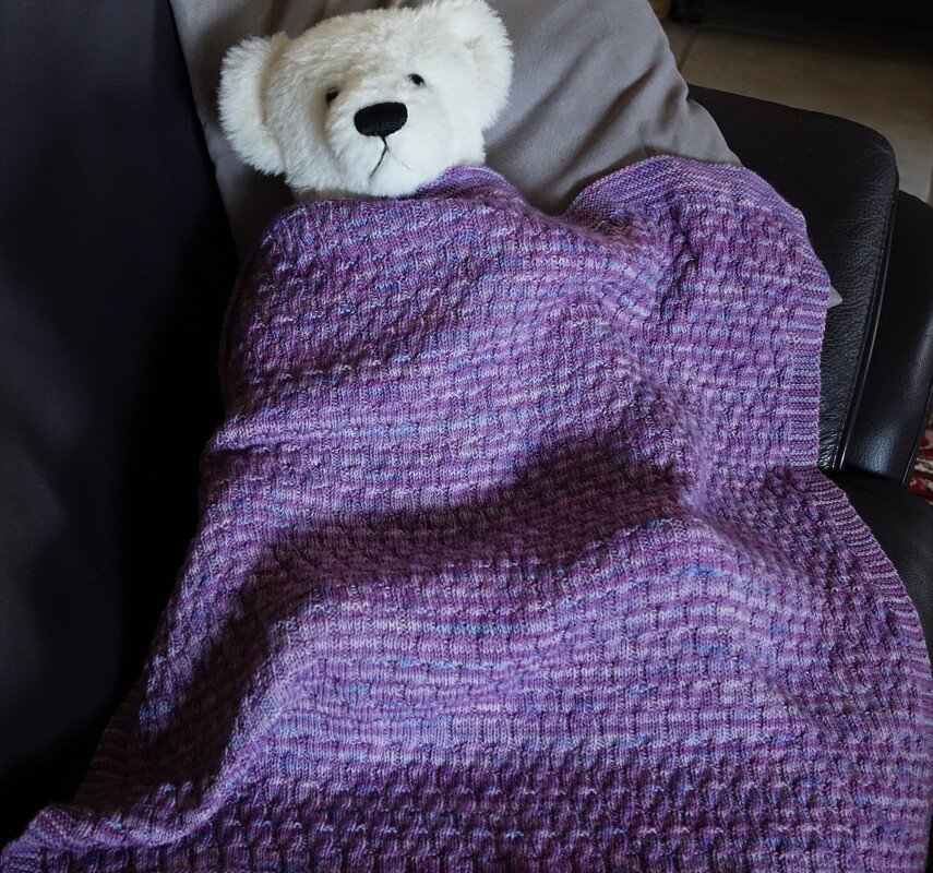 Weißer Teddybär unter lila gestrickter Babydecke mit Strukturmuster