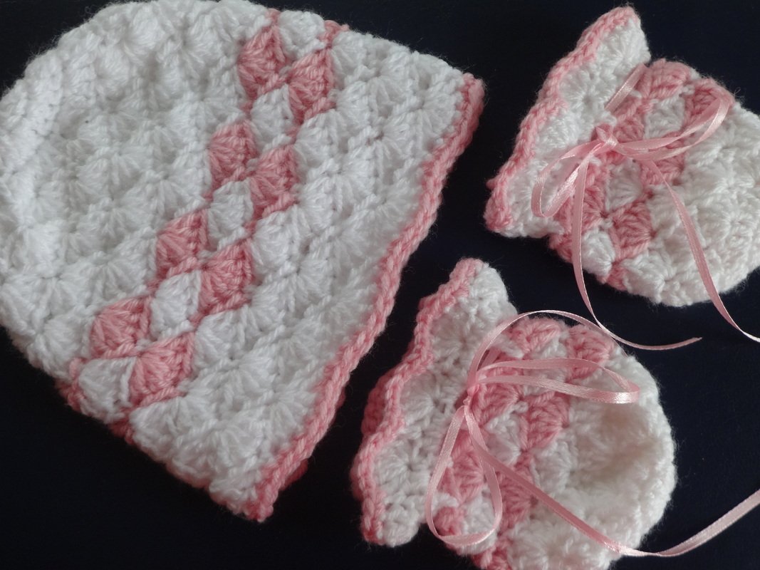 Gehäkelte weiße Mütze und zwei rosa-weiße Schühchen mit rosa Bändern auf dunklem Untergrund