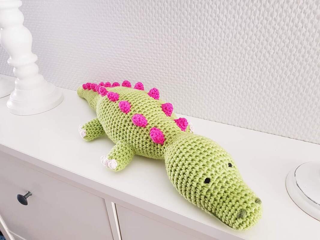 Hellgrünes gehäkeltes Amigurumi-Krokodil mit rosa Stacheln liegt auf einem weißen Sideboard