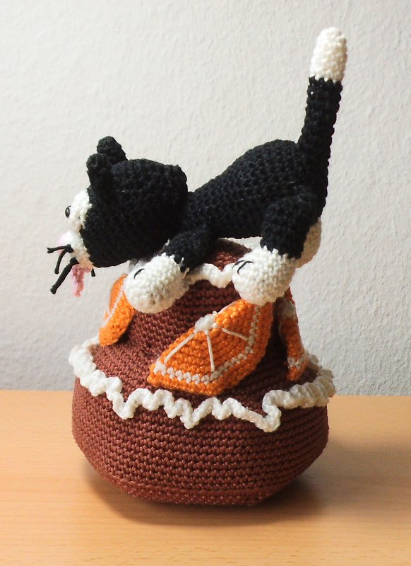 Schwarz-weiße gehäkelte Katze auf braunem Kuchen mit orangen Dekoscheiben und weißer Rüsche