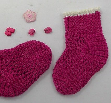 Zwei fuchsiafarbene gehäkelte Babysocken mit weißem Bündchen und rosa Dekorknöpfen auf hellem Hintergrund.