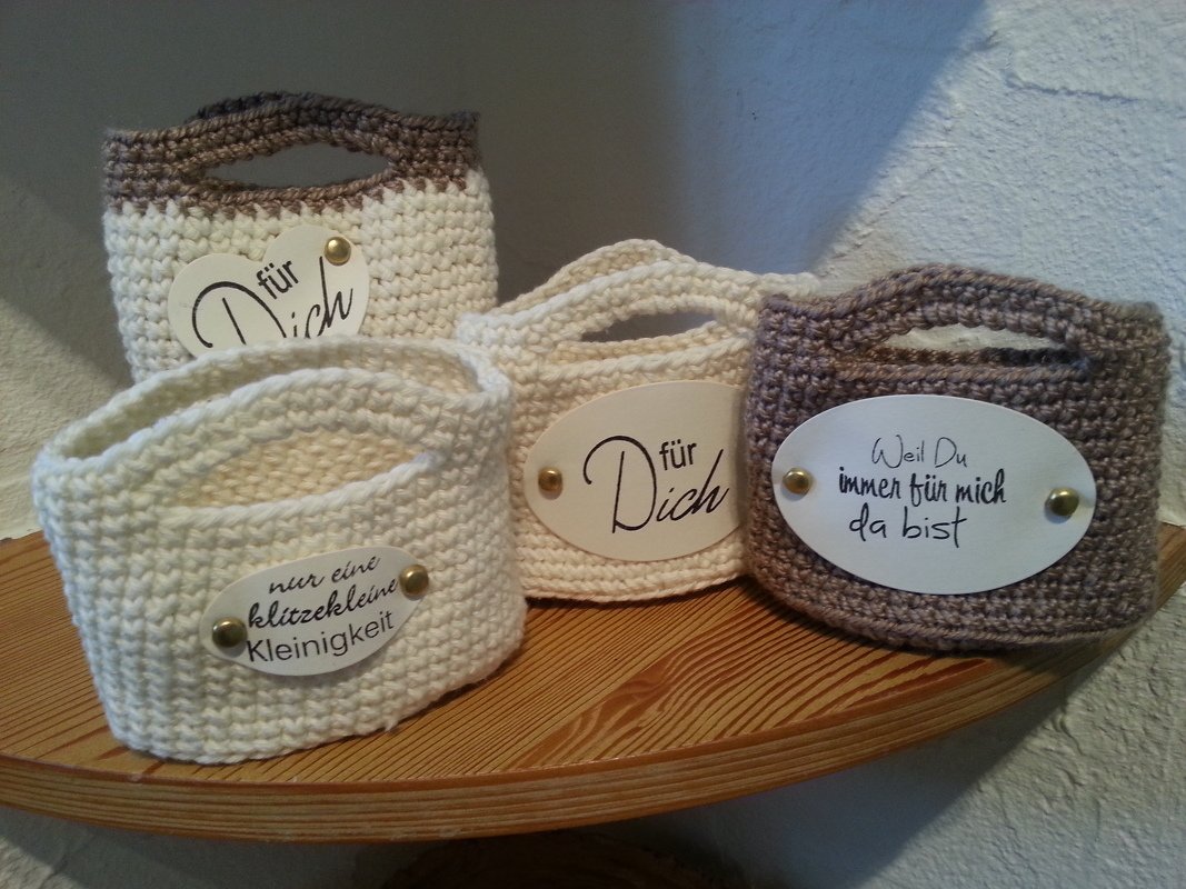 Vier gehäkelte Mini-Körbe in Creme und Braun mit Spruch-Anhängern auf einem Holzregal.