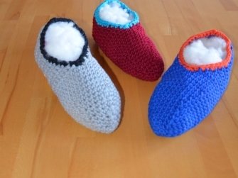 Drei gehäkelte Hausschuhe in Grau, Rot und Blau auf Holzfußboden