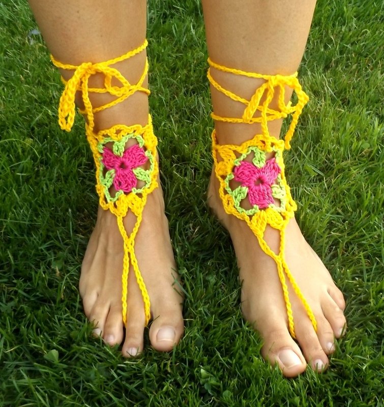 Füße mit gelben gehäkelten Barfuß-Sandalen und pink-grünem Blumenmotiv auf Gras