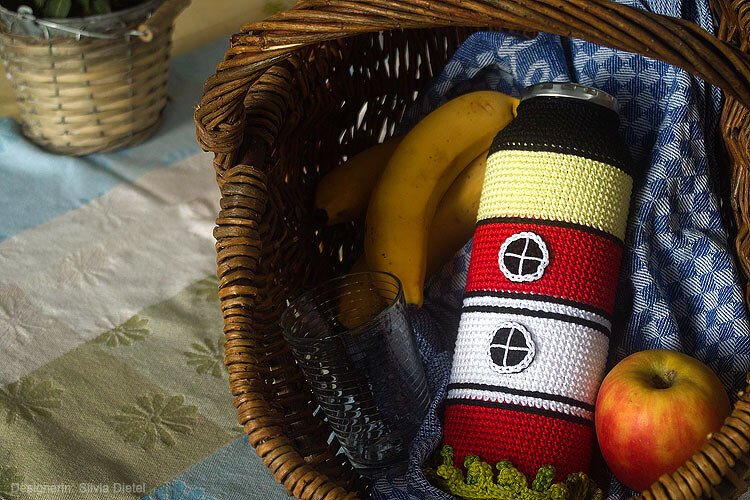 Geflochtener Korb mit gehäkelter Leuchtturm-Flaschenhülle, Banane, Apfel und Glas