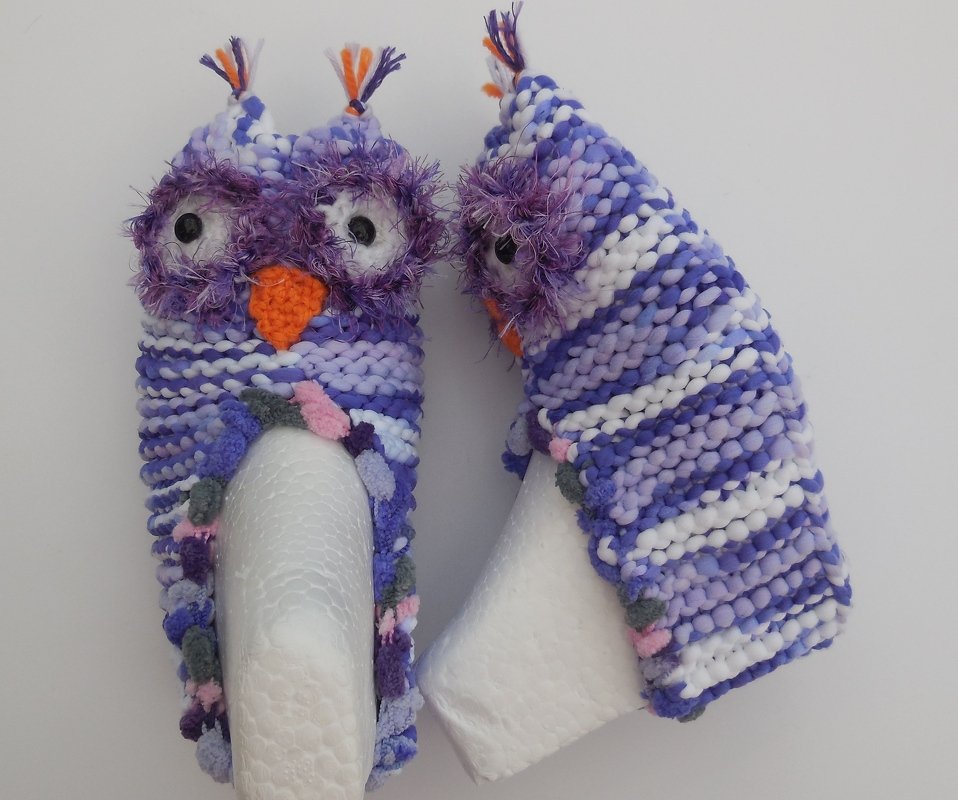Zwei gestrickte Eulen-Kinderhausschuhe in Lila und Weiß mit orangefarbenem Schnabel und flauschigen Augenrändern