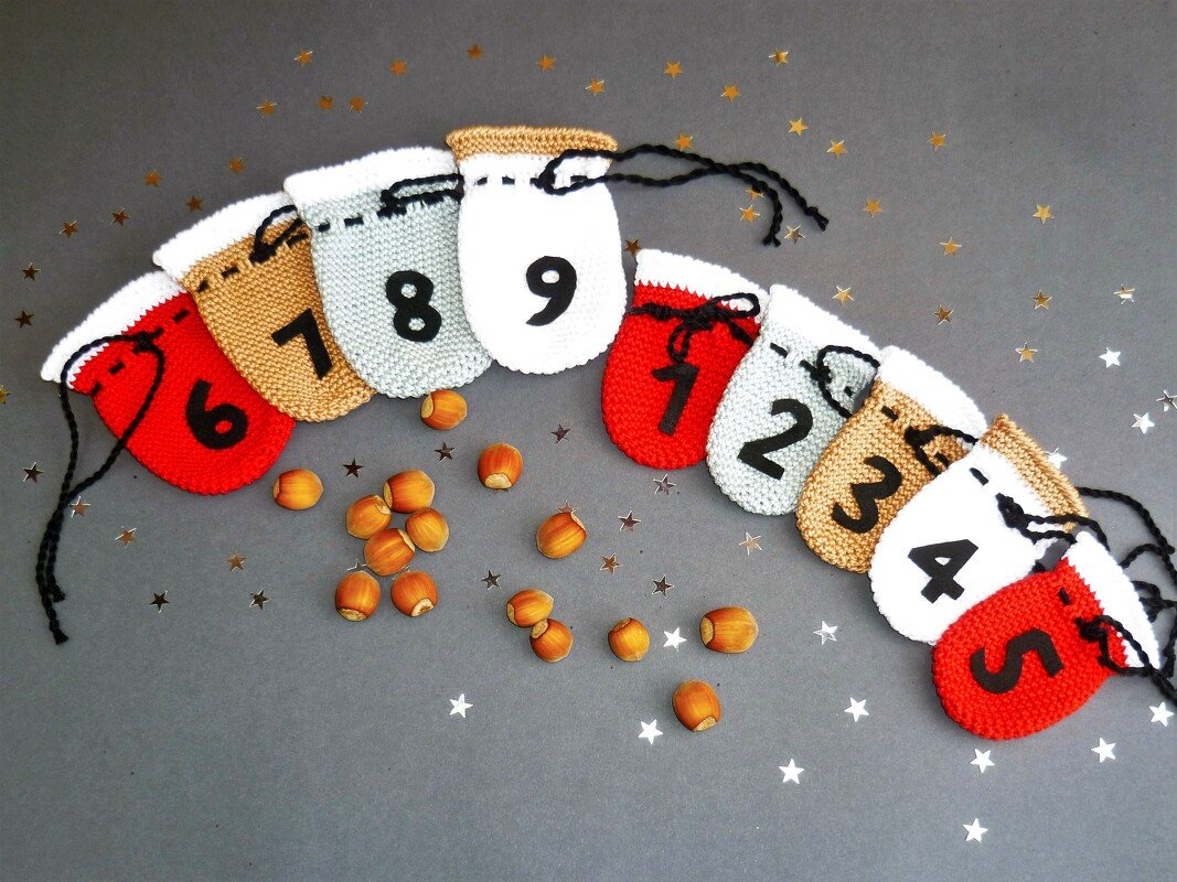 Neun gehäkelte Adventssäckchen in Rot, Hellgrau, Weiß und Braun mit schwarzen Zahlen und Kordelzügen, umgeben von Haselnüssen und Sternkonfetti.
