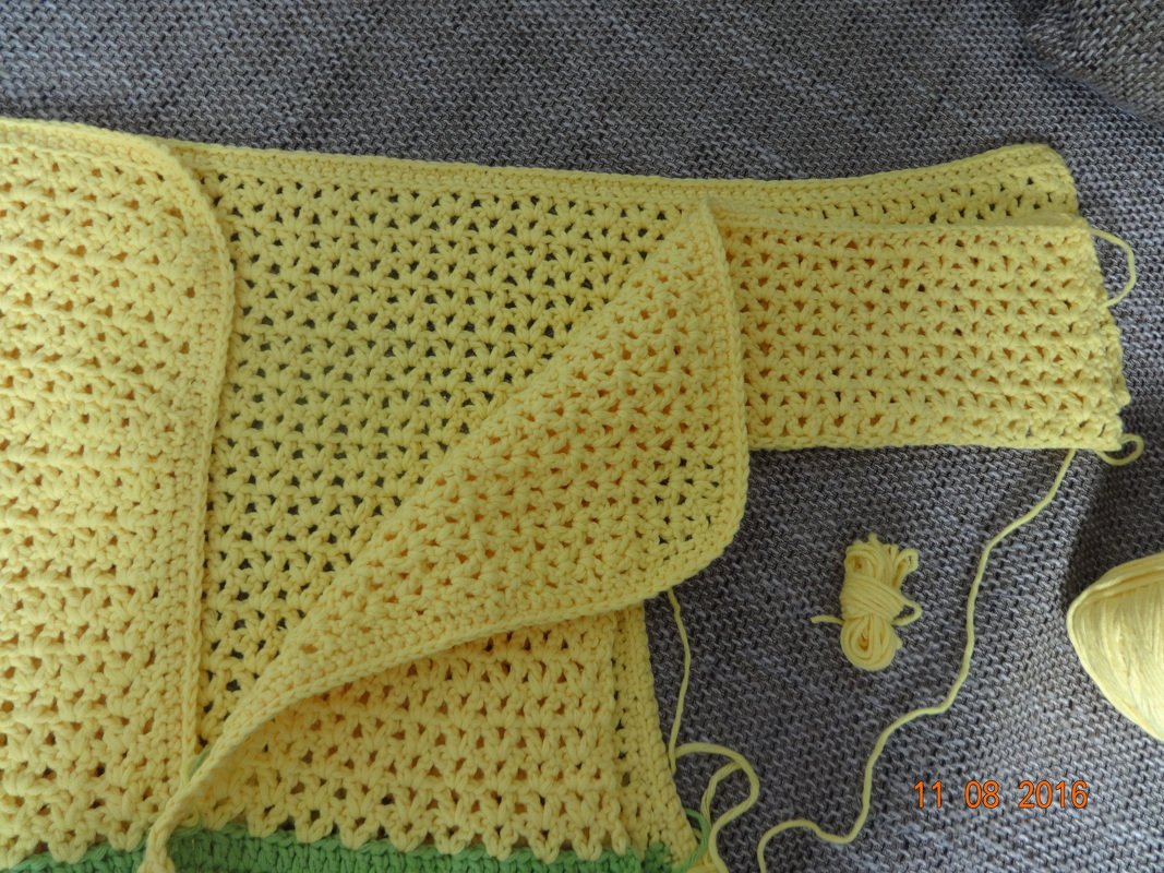 Gelbe gehäkelte Babyjacke mit V‑Stich‑Lochmuster und grünem Saum, Garnknäuel auf grauem Untergrund