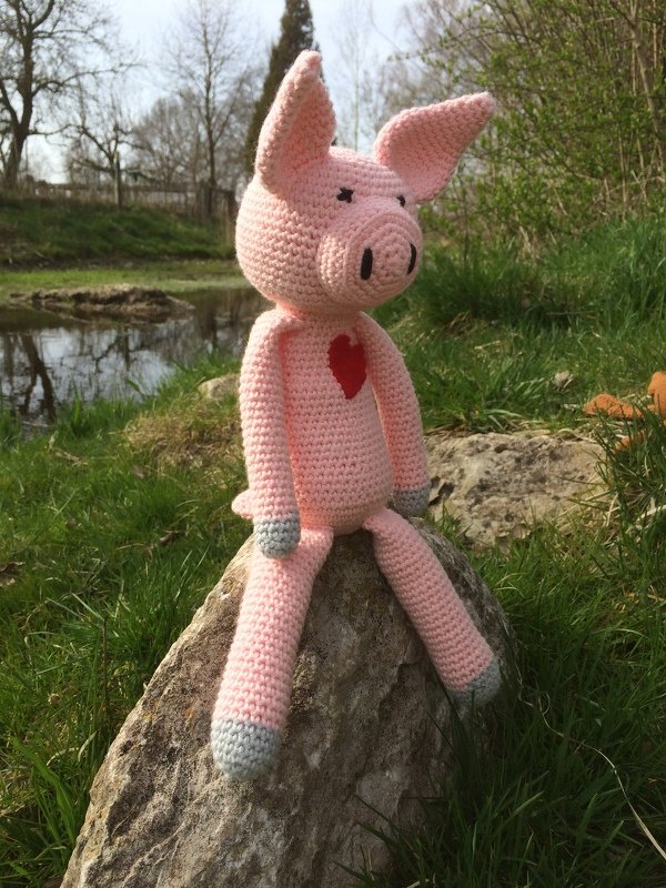 Rosafarbenes Amigurumi-Schwein mit rotem Herz und grauen Hufen sitzt auf einem Stein