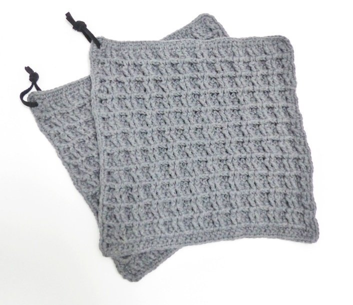 Zwei graue gehäkelte Topflappen mit Waffelmuster und schwarzen Aufhängeschlaufen auf hellem Hintergrund.