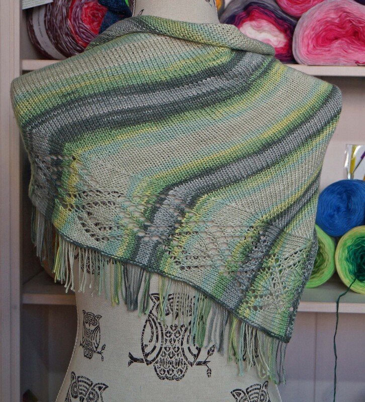 Gestricktes Dreieckstuch in Grün, Grau und Gelb mit Streifen, Lochmusterborte und Fransen auf einer Schneiderpuppe vor Garnregal.