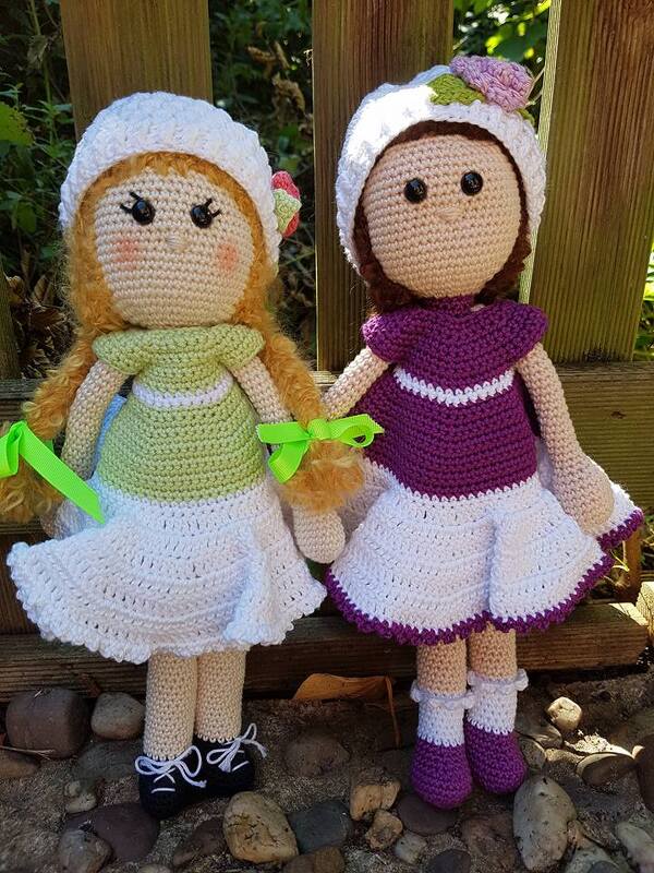Zwei gehäkelte Amigurumi-Puppen mit weißen Röcken, Oberteilen in Grün und Lila, Mützen mit Blüte und Schuhen vor einem Holzzaun