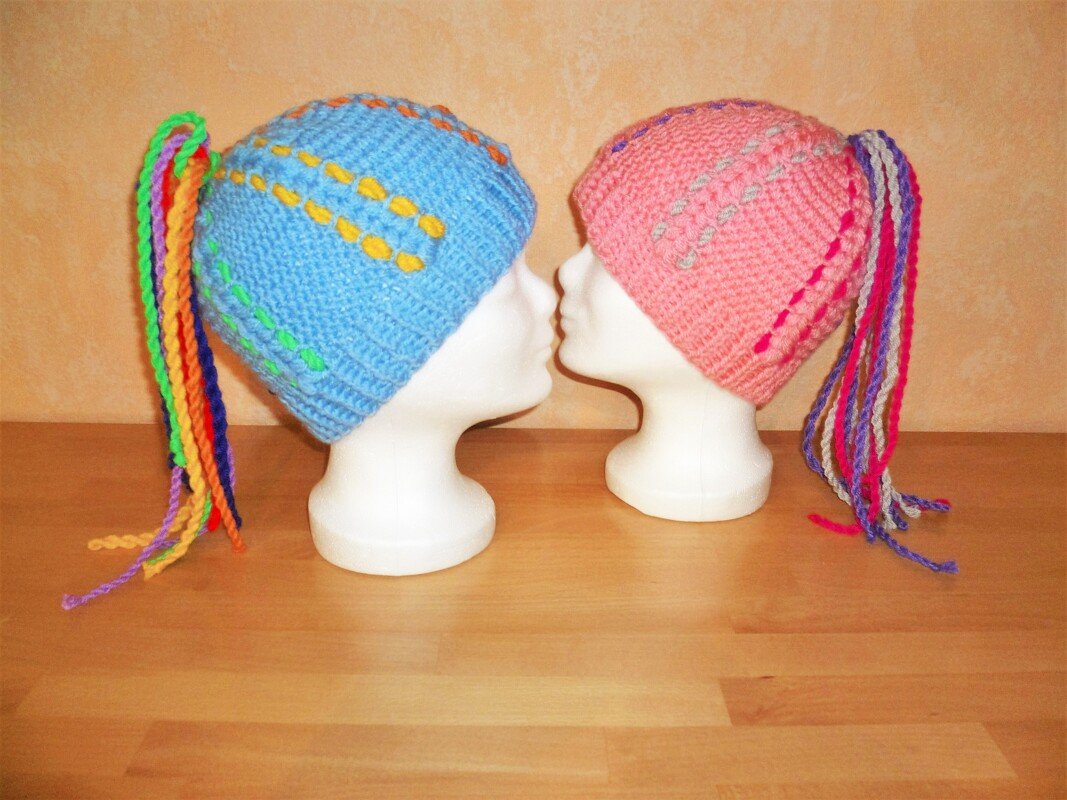 Gehäkelte blaue und rosa Mütze mit bunten Zöpfen auf weißen Kopfformen