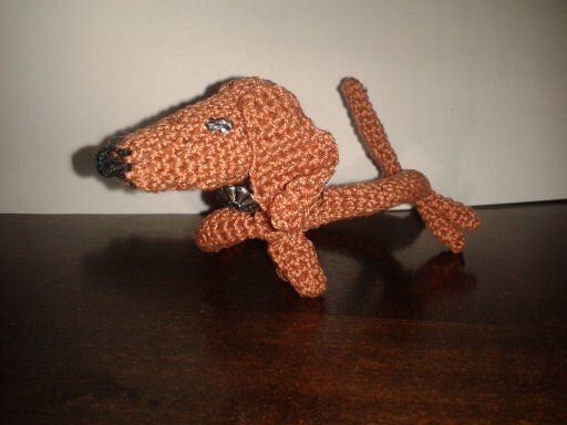 Braunes gehäkeltes Dackel-Amigurumi mit Nieten-Halsband auf dunkler Holzfläche.