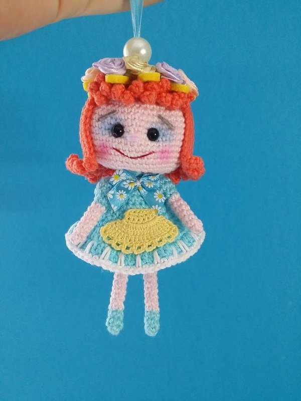 Gehäkelte Amigurumi-Puppe mit roten Haaren, hellblauem Gänseblümchenkleid, gelber Schürze und Blütenkranz, aufgehängt an Perle vor blauem Hintergrund.