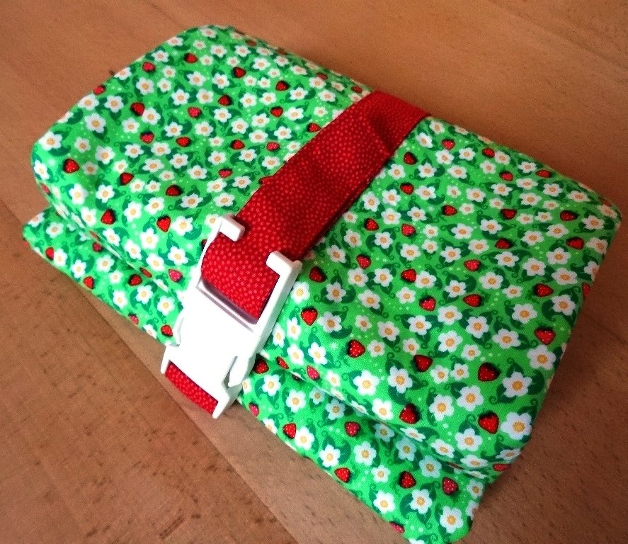 Gerolltes Utensilo aus grünem Stoff mit Erdbeeren und Gänseblümchen, rotes Gurtband und weiße Schnalle