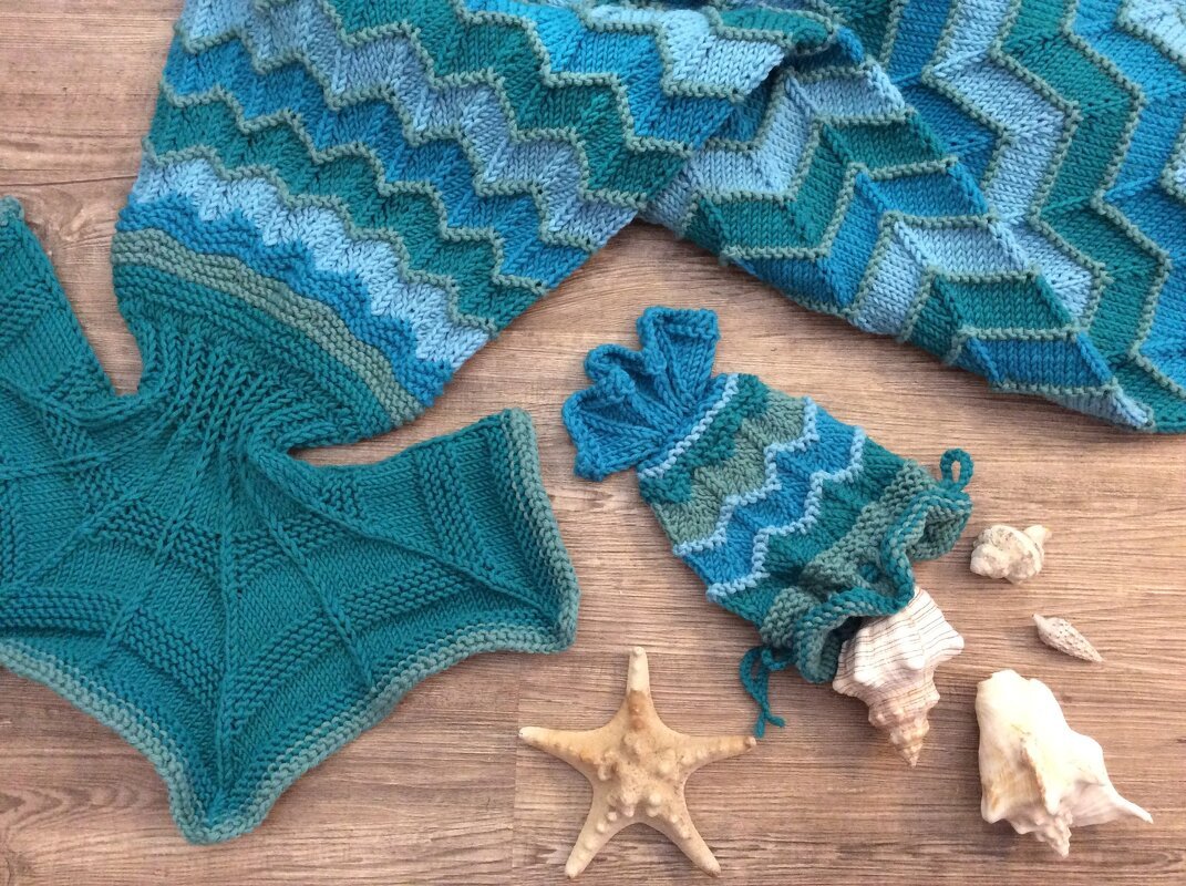 Handgestrickte Chevron-Decke, Meerjungfrauenflosse und Kordelzugbeutel in Türkis- und Blautönen auf Holz mit Muscheln und Seestern.