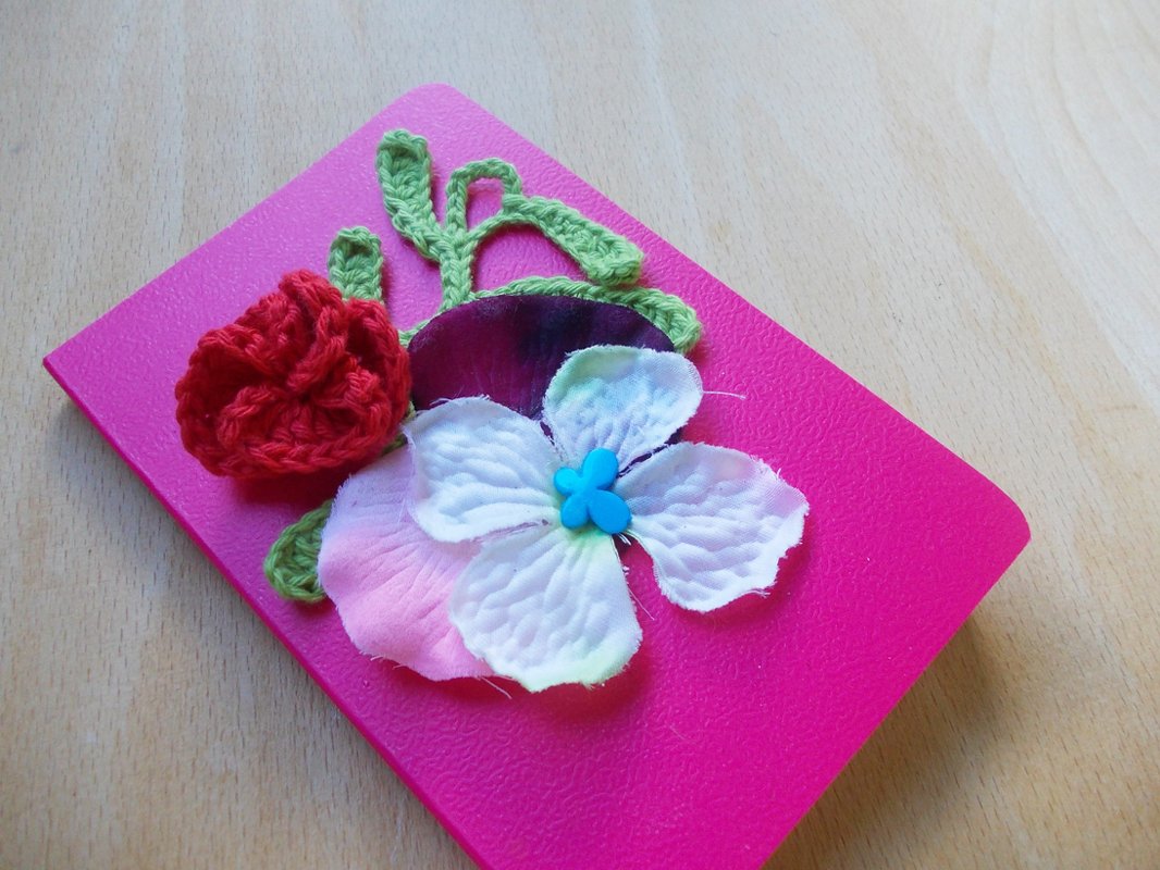 Pinkes Notizbuch mit roter Häkelrose, weiß-rosafarbener Stoffblüte und grünen Häkelblättern