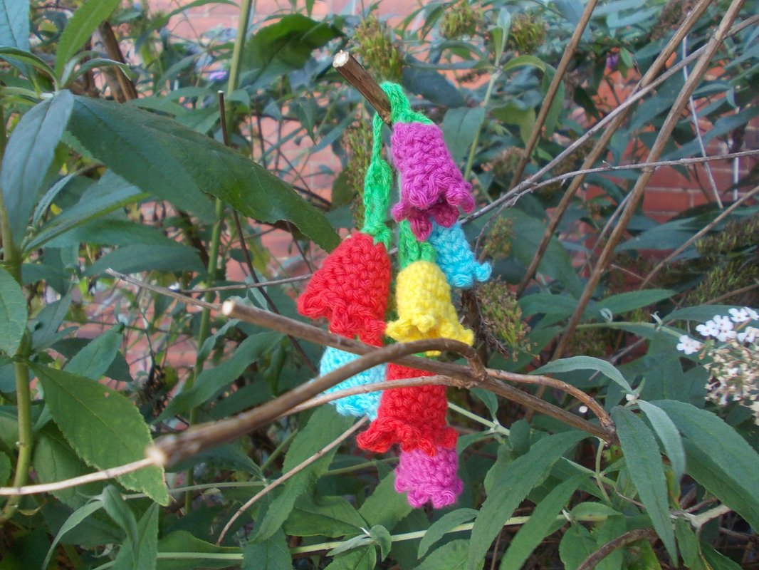 Nahaufnahme bunter gehäkelter Glockenblumen an einem Ast vor grünen Blättern.