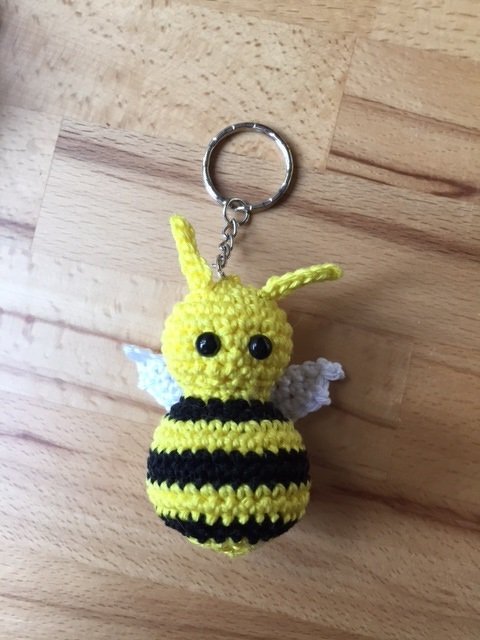 Gehäkelter Bienen-Schlüsselanhänger mit gelb-schwarzen Streifen, weißen Flügeln und Metallring auf Holz.